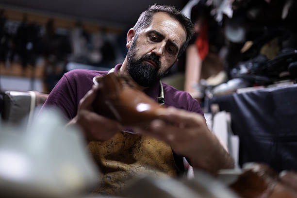 A skilled craftsman with a beard and apron carefully shapes a leather shoe in a busy workshop filled with tools and materials, highlighting traditional leatherwork and focus.