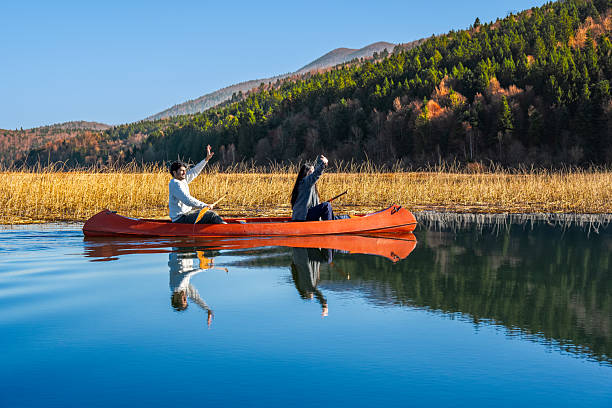 Two individuals paddle a red canoe on a serene lake, surrounded by autumnal forests. Their reflections ripple in the calm water, capturing a moment of tranquility and connection with nature. The scene evokes themes of adventure and peaceful exploration in a picturesque outdoor setting.
