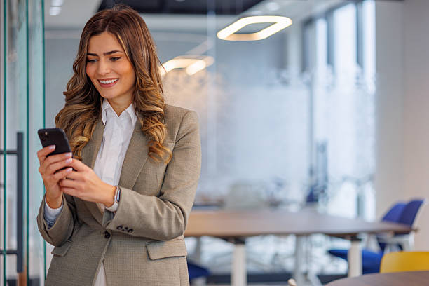 istock Smiling businesswoman standing in an open plan office, using a smartphone for communication, networking, and business technology 2253431894