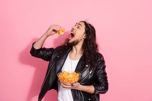 istock Bold nonbinary LGBT man in leather jacket and tiara enjoys chips against pink pastel background, 2253435251