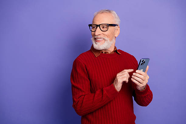 istock Mature elderly man in red sweater smiles while using a smartphone against a purple background for lifestyle business advertising and fashion branding 2253436001