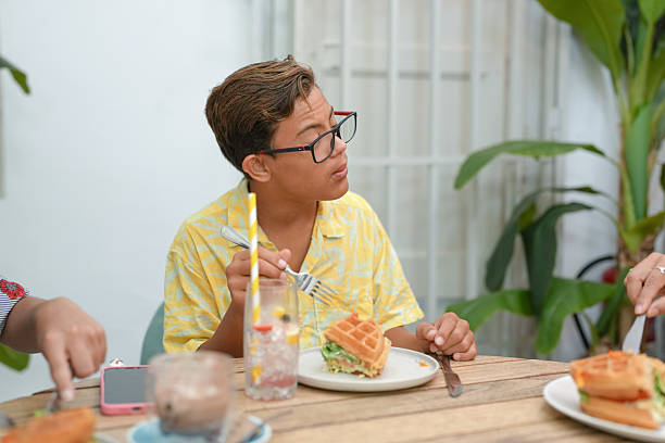 A young man in glasses is eating a waffle sandwich. He is sitting at a table with a fork and knife in front of him. There are other people in the background, and a potted plant is visible