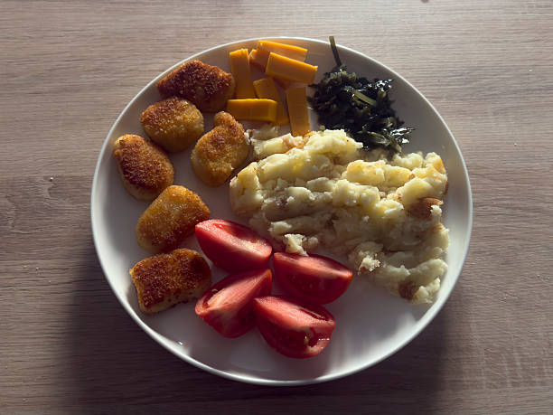 A plate holds chicken nuggets, mashed potatoes, cheese, tomatoes, and greens at home.