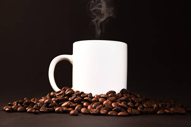 Steaming hot coffee in a white mug amid scattered roasted beans on a dark rustic wooden surface, warm and aromatic ambiance
