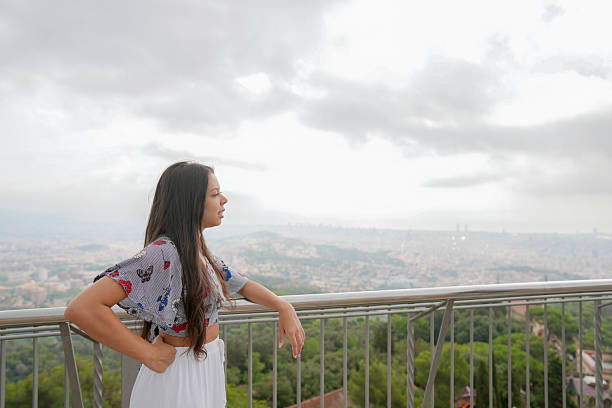 A woman stands on a balcony overlooking a city. The sky is cloudy and the woman is looking out over the city