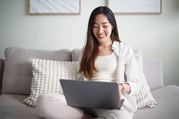 istock Smiling asian woman sitting on sofa using laptop for remote work or online learning at home. 2253449660
