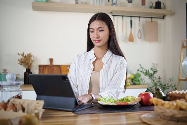 Smiling young woman using digital tablet while eating a healthy breakfast with salad and fruits in a cozy kitchen.