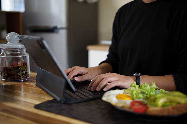 Young woman working on digital tablet in the kitchen while having a healthy breakfast with eggs, salad, and avocado toast.