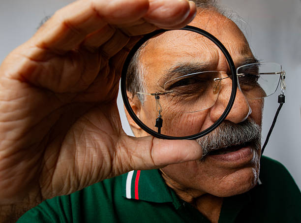 Close-up suspicious senior detective in eyeglasses looking through magnifying glass against white background