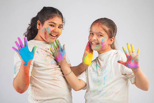Portrait of playful sisters applying colorful powders on each other faces while celebrating Holi together on white background