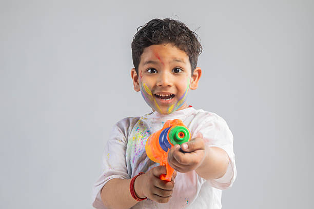 Portrait of cute playful boy with color powder on face aiming squirt gun against white background