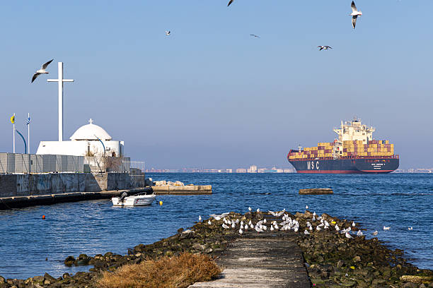 Thessaloniki, Greece – 14 December 2025: A large MSC container ship sails through the Thermaic Gulf near the waterfront of Thessaloniki, with a small seaside Orthodox chapel and seagulls in the foreground. The scene highlights everyday maritime activity, global trade routes, and the close relationship between the port infrastructure and urban coastal life in northern Greece.