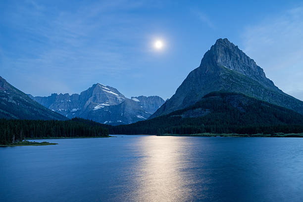 A full moon rises over the dramatic peaks of Glacier National Park in Montana, casting a golden reflection across the Swiftcurrent Lake. Majestic mountains and dense forest complete this serene and captivating wilderness scene.