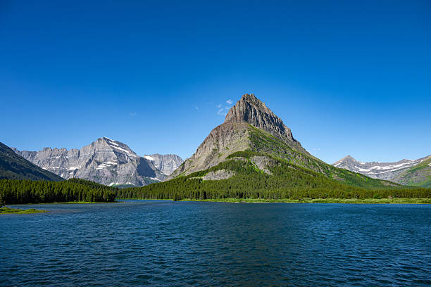 Towering mountain peaks rise above a still alpine lake in Glacier National Park, Montana. The surrounding evergreen forest and vibrant blue sky create a timeless scene of natural tranquility and rugged wilderness.