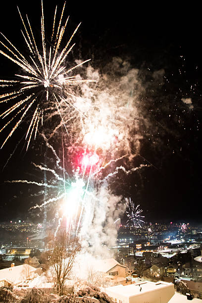 Spectacular New Year's Eve celebration in Trondheim, Norway. Colorful fireworks and faint Aurora Borealis illuminate the night sky over snowy houses, with spectators watching the display from a viewpoint.