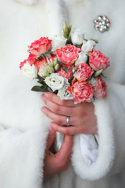 Close-up of a woman's hand in a white fur coat gently holding a vibrant bouquet of pink carnations and white roses, featuring a silver ring on her finger and a sparkling brooch in the soft background, evoking romance and wedding elegance.