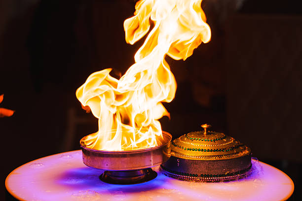 Bright flames surge from a metal bowl, creating an intense display of fire. The warm glow highlights the details in the dark surrounding environment.