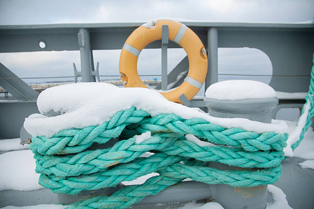 A close-up shot of a heavy turquoise mooring rope covered in fresh snow, wrapped around a bollard on a ship's deck, with an orange lifebuoy in the blurred background.
