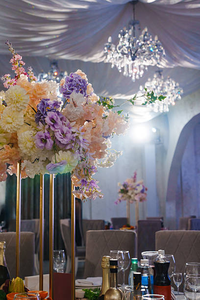 Colorful flower centerpieces stand tall in a beautifully decorated banquet hall. Soft lighting creates a warm and inviting atmosphere for guests.