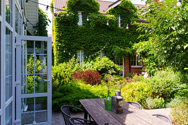 A garden features various plants and shrubs with a wooden table. Sunlight shines down, and string lights hang above. The space is suitable for gatherings or meals.