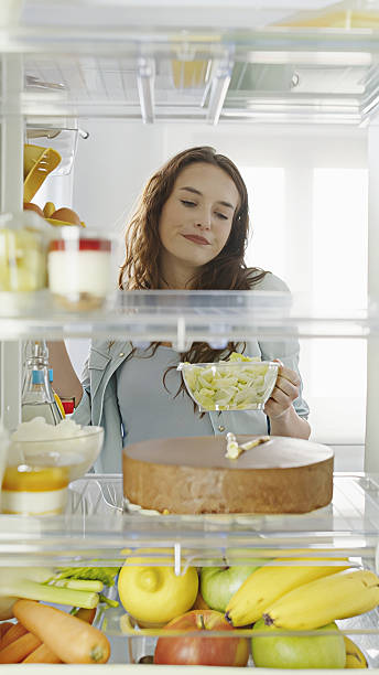 istock Woman stands in front of open fridge, reluctantly deciding between a dessert and a fresh salad. She chooses the healthy option, making a healthy diet choice. POV shot from inside kitchen refrigerator 2253473298