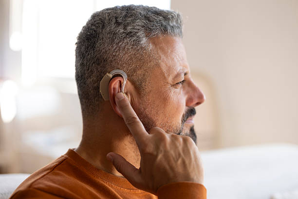 Mid adult man adjusting his hearing aid at home, using assistive technology to support communication and independent lifestyle.