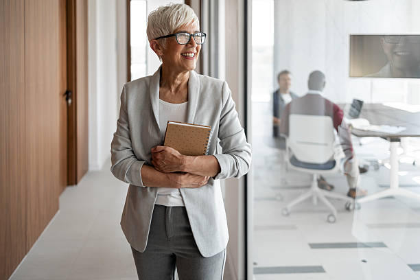 istock Confident Senior Professional Smiling In Modern Office Hallway With Notebook 2253476183