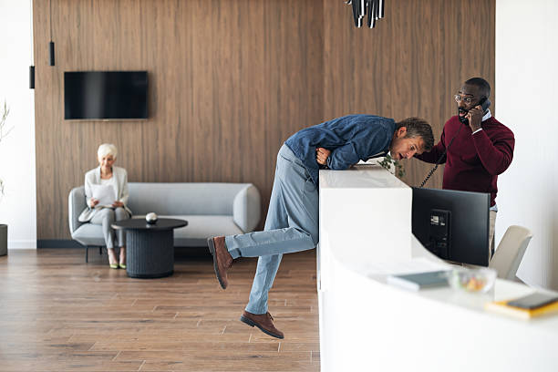 A modern office lobby shows a man leaning over the reception counter on a phone, another staff member assisting, and an elderly woman waiting on a sofa. A candid, professional workplace moment.