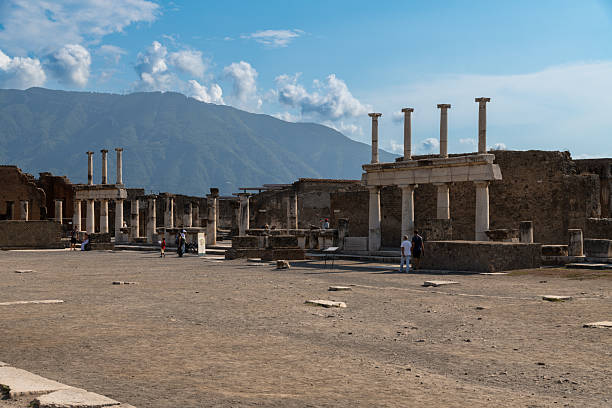 Pompei â Campania â Italy â 08-25-2025: Tourists walk across the ancient Forum in Pompeii, surrounded by stone ruins and columns, with a mountain ridge under a bright blue sky.