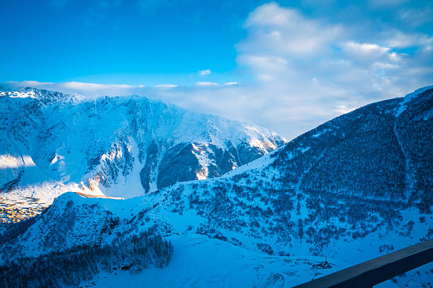 A high viewpoint from Gergeti Holy Trinity Church looks across steep snowcovered ridges to sunlit cliffs and a bright blue sky with soft clouds in the Caucasus Mountains.