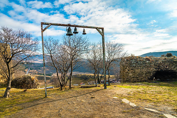 A simple metal frame holding three church bells stands among bare trees and stone ruins near Jvari Monastery, overlooking rolling hills under a bright blue sky with soft clouds.