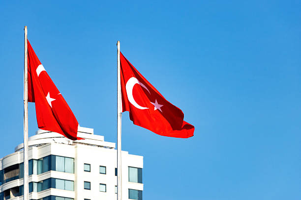 Two Turkish flags flying on flagpoles against clear blue sky—framed by modern white building with balconies in a clean architectural scene evoking national pride and visual contrast.