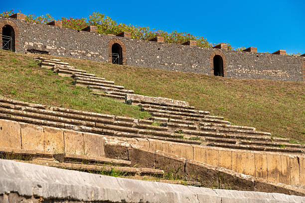 Pompei â Campania â Italy â 08-25-2025: Terraced stone seating on a grassy slope in the Amphitheater Pompeii, with arched entrances in a lava stone wall under clear blue sky.
