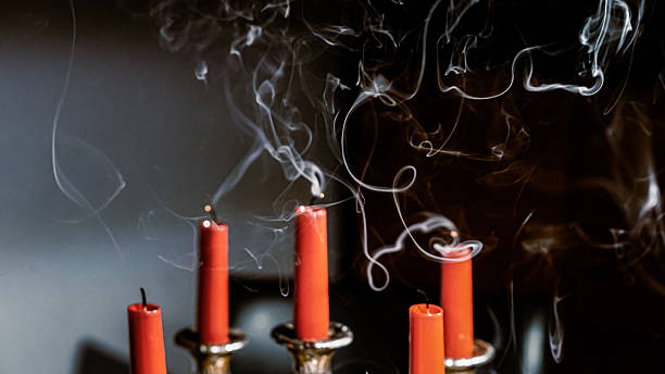 Minimalist close-up of five red candles recently extinguished, with delicate trails of smoke rising from each wick. The candles are placed in a silver holder against a dark background. The swirling smoke contrasts with the stillness of the scene, evoking themes of transience, silence, and visual simplicity