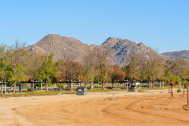 Mountains rise over Lake Perris State Recreation Area's picnic grounds and dusty terrain, with trees showing autumn colors under clear blue sky.