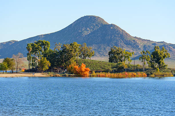 Lake Perris State Recreation Area's tranquil waters reflect the clear sky, with vibrant autumn foliage along the shore and a rugged mountain beyond.
