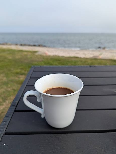 a Close-up of a white mug filled with coffee standing on a wooden table in the garden overlooking the sea