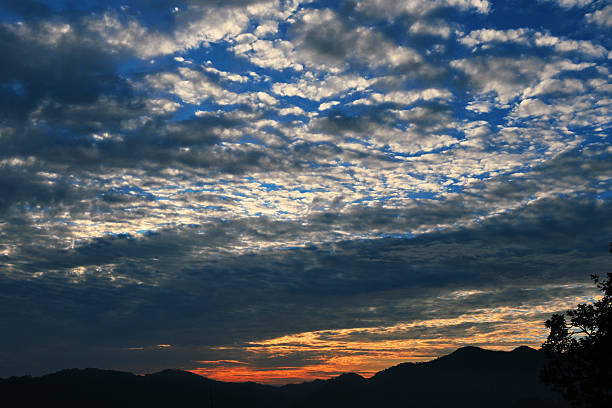 A striking evening sky filled with textured clouds glowing in shades of blue, gold, and orange above dark mountain silhouettes. The scene captures the calm yet dramatic transition from day to night, evoking moods of serenity, wonder, and reflection. Ideal for themes related to nature, weather, landscapes, tranquility, travel, background skies, and atmospheric storytelling.