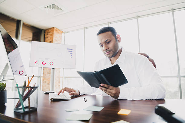 istock Young handsome entrepreneur reviews document in modern loft office with charts and keyboard at desk 2253499131