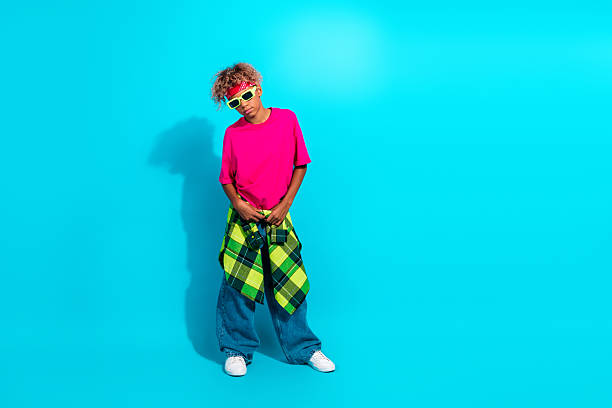 A young boy with curly hair wears a bright pink shirt and baggy jeans with a green plaid shirt tied around the waist, posing against a vivid blue backdrop for a playful street style look.