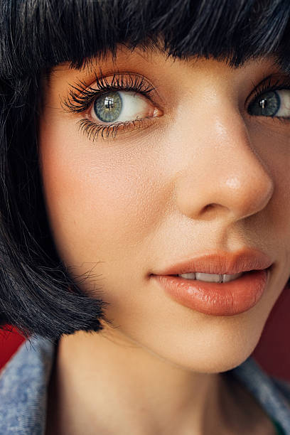 An intimate close-up beauty portrait focusing on expressive eyes, delicate skin texture and soft natural makeup. The low-angle framing highlights the long eyelashes, smooth complexion and warm peach tones on the lips and cheeks. The short black bob haircut frames the face with graphic precision, while the subtle background color adds depth without distraction. The image conveys freshness, youth, and an effortless modern beauty ideal suitable for skincare, cosmetics, fashion and editorial storytelling.