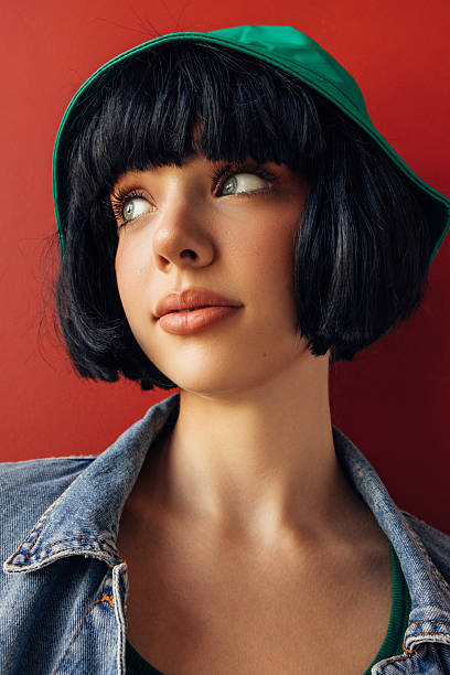 A stylish close-up portrait of a young woman with a short black bob haircut and a green cap, captured in a calm reflective moment against a rich red background. Her gaze turned slightly away from the camera creates a sense of inner dialogue and quiet curiosity. Soft natural light highlights the smooth texture of her skin, subtle makeup and delicate facial features. The contrast between the green cap, denim jacket and warm red backdrop builds a modern editorial mood suitable for fashion, lifestyle and beauty storytelling.
