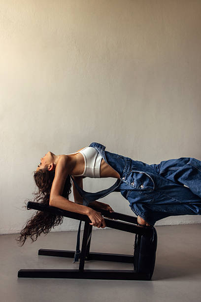 A woman in a white sports bra and denim jacket bends backwards over a sturdy bench in a minimal studio. The pose highlights flexibility, balance, and athleticism, with natural light and a calm, focused mood. Suitable for fitness, wellness, or fashion-related stock highlights.