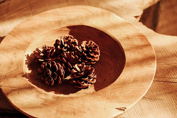 Four pine cones sit in a wooden bowl on a table. Sunlight shines on the bowl creating warm tones. The setting shows a natural look with wood textures and the beauty of simple items.