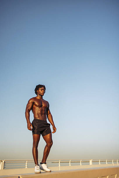 A muscular man in black shorts and white sneakers stands confidently on a seaside boardwalk, bathed in golden hour light. The expansive blue sky and calm horizon emphasize fitness, strength, and freedom. This image suits sports, lifestyle, and wellness campaigns with a bold, active vibe.
