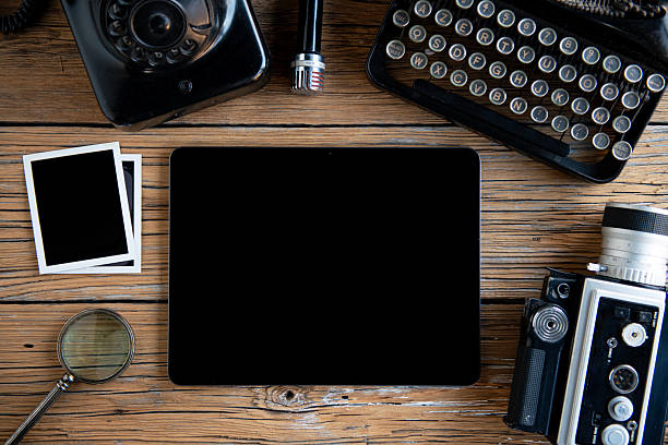 Top view flat lay of a creative workspace combining vintage equipment and modern technology. Retro typewriter, analog camera, old telephone and classic microphone arranged around a modern tablet with blank screen on rustic wooden table. Concept of contrast between old and new, creativity, content creation, storytelling and copy space background.