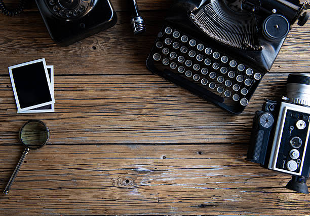 Top view flat lay of a vintage creative workspace with retro typewriter, analog camera, old telephone, classic microphone, magnifying glass and blank instant photo frames on rustic wooden table. Nostalgic background with copy space for text, design, storytelling, journalism and creative concepts.