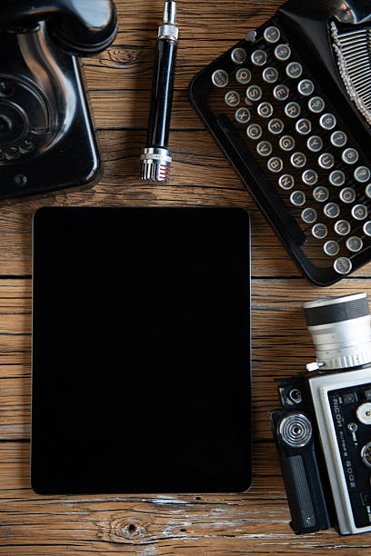 Top view flat lay of a creative workspace combining vintage equipment and modern technology. Retro typewriter, analog camera, old telephone and classic microphone arranged around a modern tablet with blank screen on rustic wooden table. Concept of contrast between old and new, creativity, content creation, storytelling and copy space background.