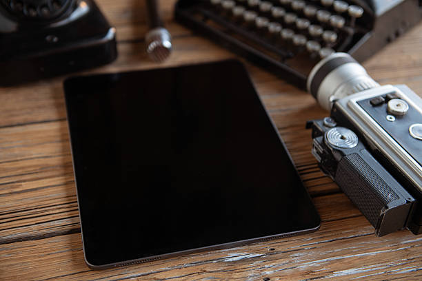 Top view flat lay of a creative workspace combining vintage equipment and modern technology. Retro typewriter, analog camera, old telephone and classic microphone arranged around a modern tablet with blank screen on rustic wooden table. Concept of contrast between old and new, creativity, content creation, storytelling and copy space background.