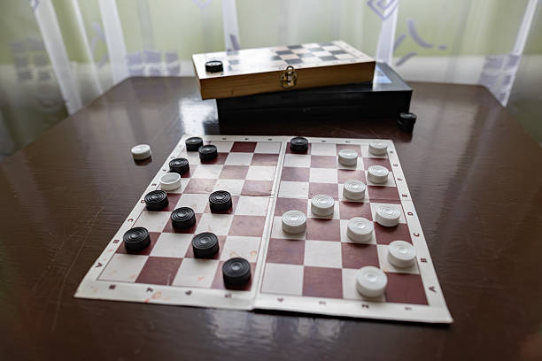 Wooden checkers board with white and black pieces on a table. Traditional indoor strategy game for leisure and mental activity. Selective focus on gaming pieces in home or club interior vibe.